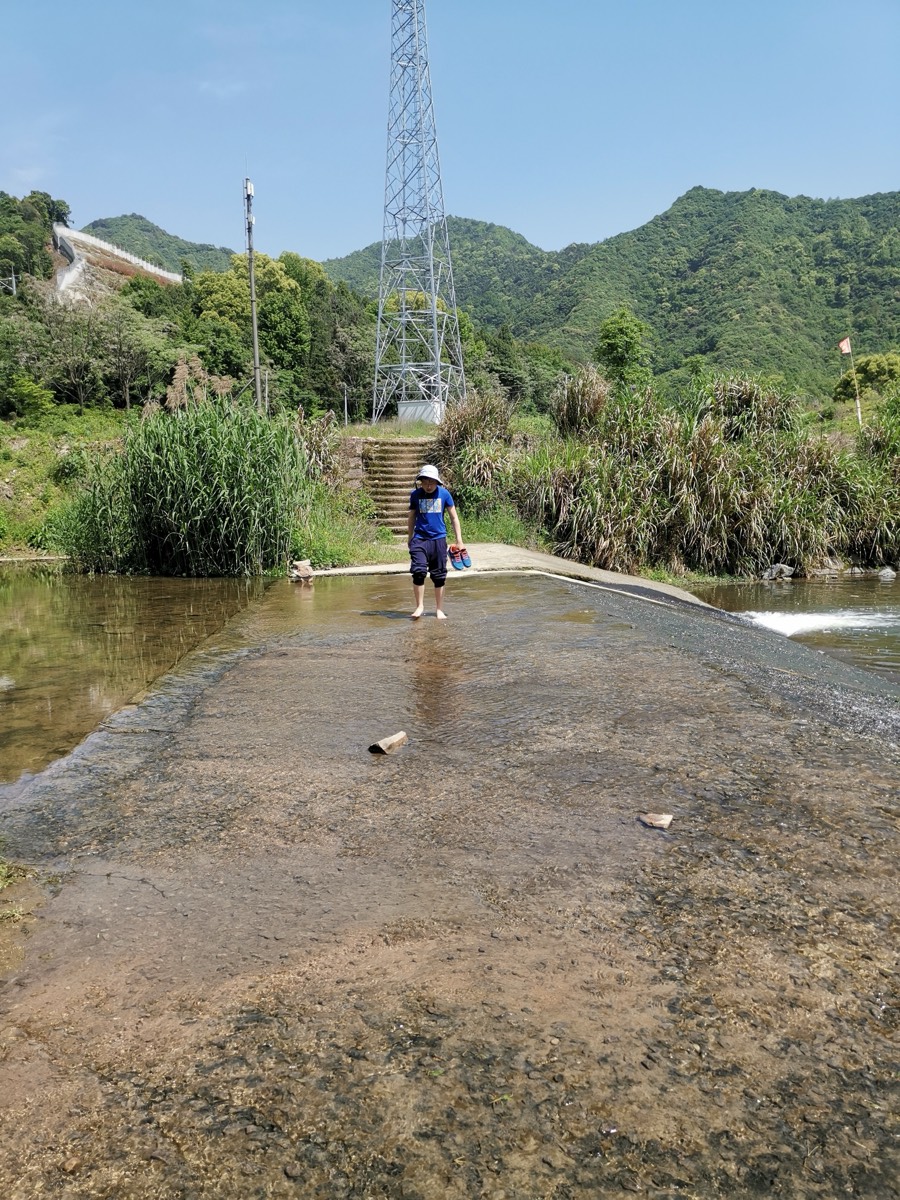 一个人提鞋赤脚站在浅浅的河面道路中央,远处青山与输电铁塔